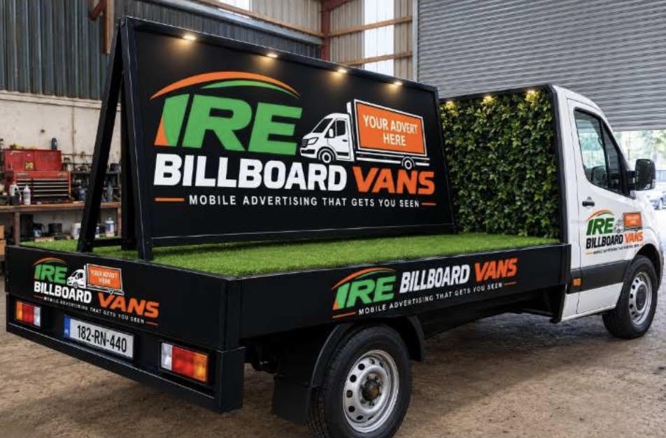IRE Billboard Vans flatbed van with a lit A-frame billboard on a grass-covered bed, branded side panels and hedge wall behind the cab